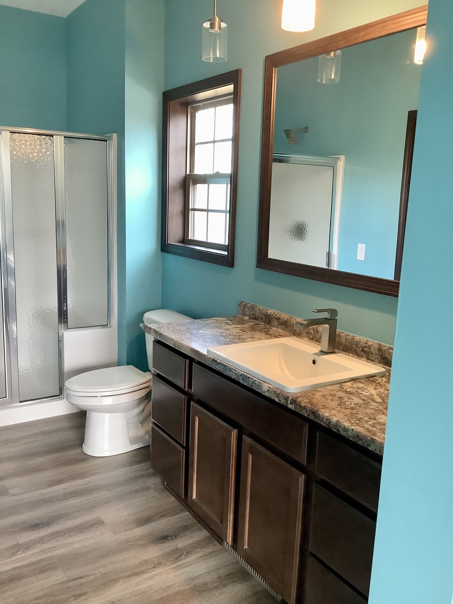 A modern bathroom with turquoise walls, wood-look flooring, and a granite countertop. Features a large mirror, sink, toilet, and glass shower door.