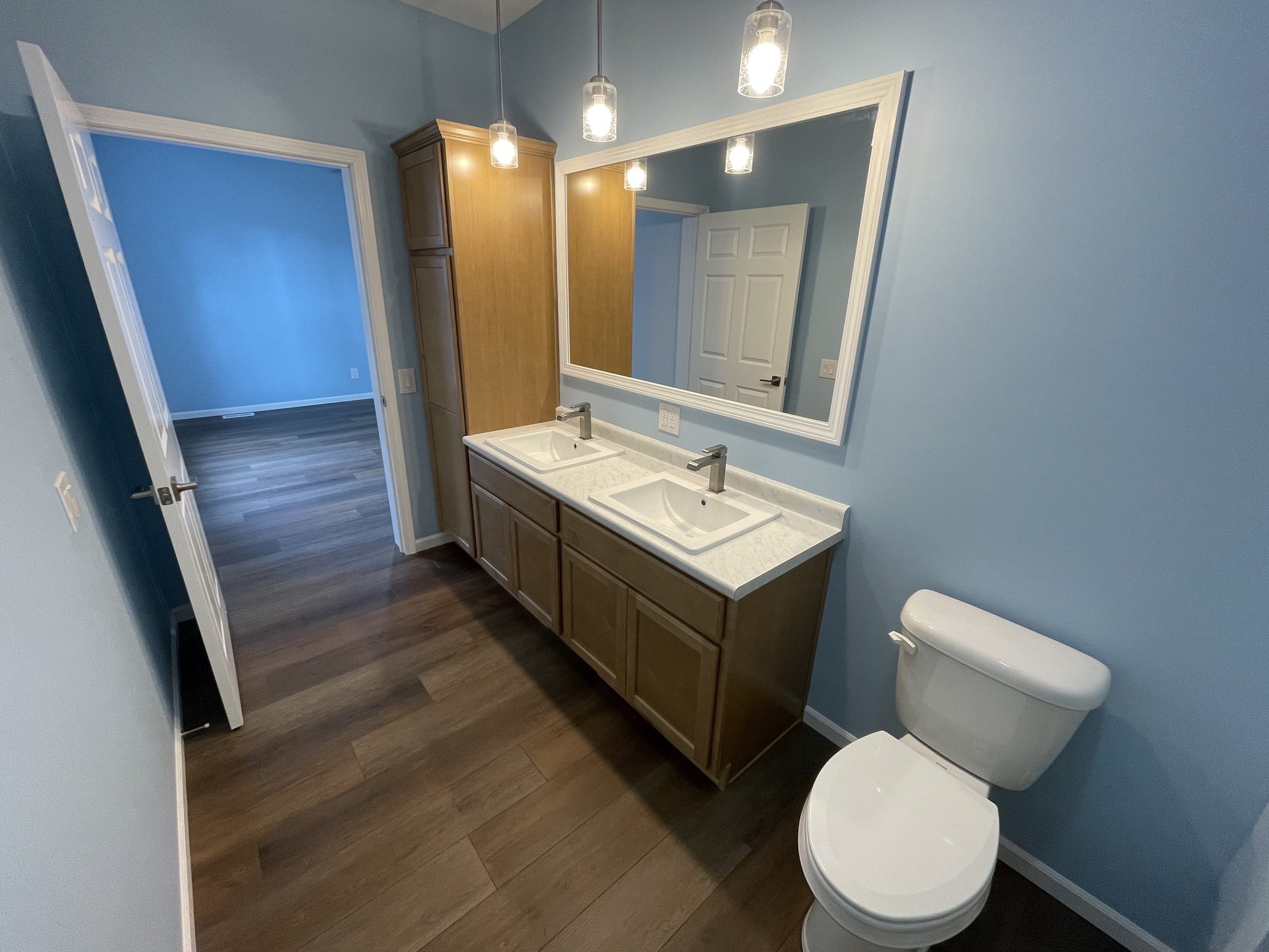 Bathroom with blue walls, dual sinks with wooden cabinets, large mirror, and a white toilet. Three pendant lights add a warm, inviting glow.