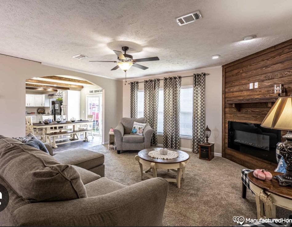 Spacious living room with beige sofas and armchair, wooden accents, and a cozy fireplace. Dining area visible through archway. Warm, inviting atmosphere.