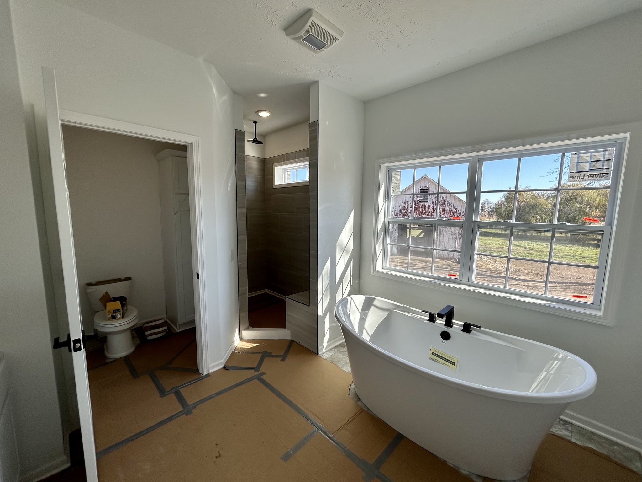 Bright bathroom under renovation, featuring a freestanding bathtub, large window with outdoor view, walk-in shower, and a partially visible toilet.
