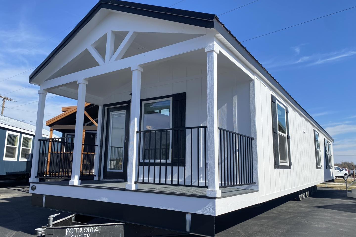 Mobile home with white siding and black trim, featuring a front porch with black railings. Clear blue sky in the background, suggesting a sunny day.