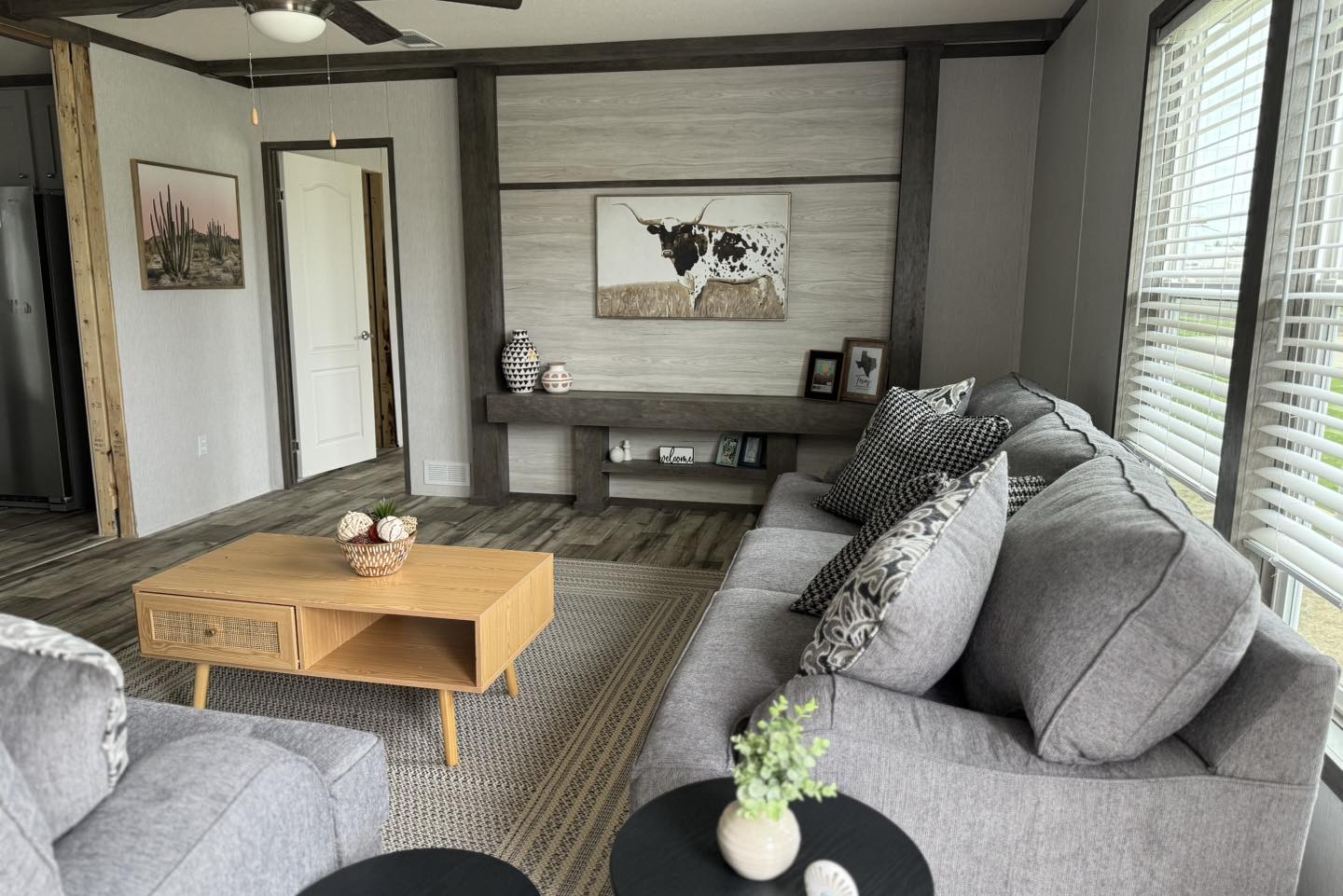 Modern living room with gray sofas, a wooden coffee table, and decorative pillows. A large window allows natural light. A cow painting and vases adorn the walls.