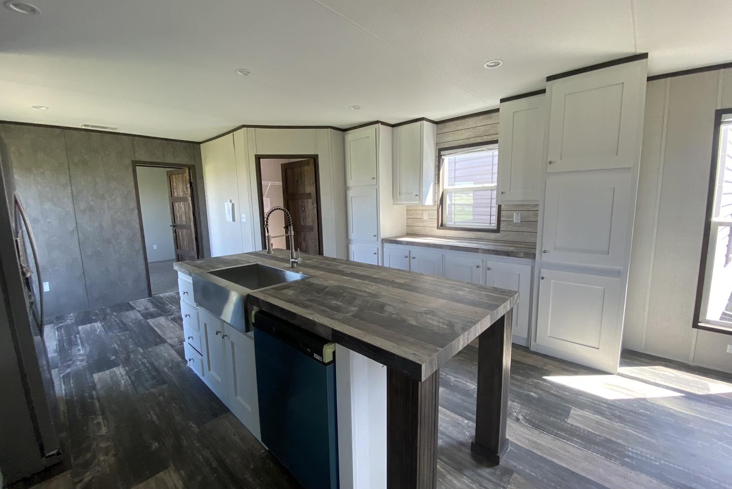 Modern kitchen with an island featuring a sink and dishwasher, wooden flooring, white cabinets, and a sunlit window, creating a bright and airy feel.