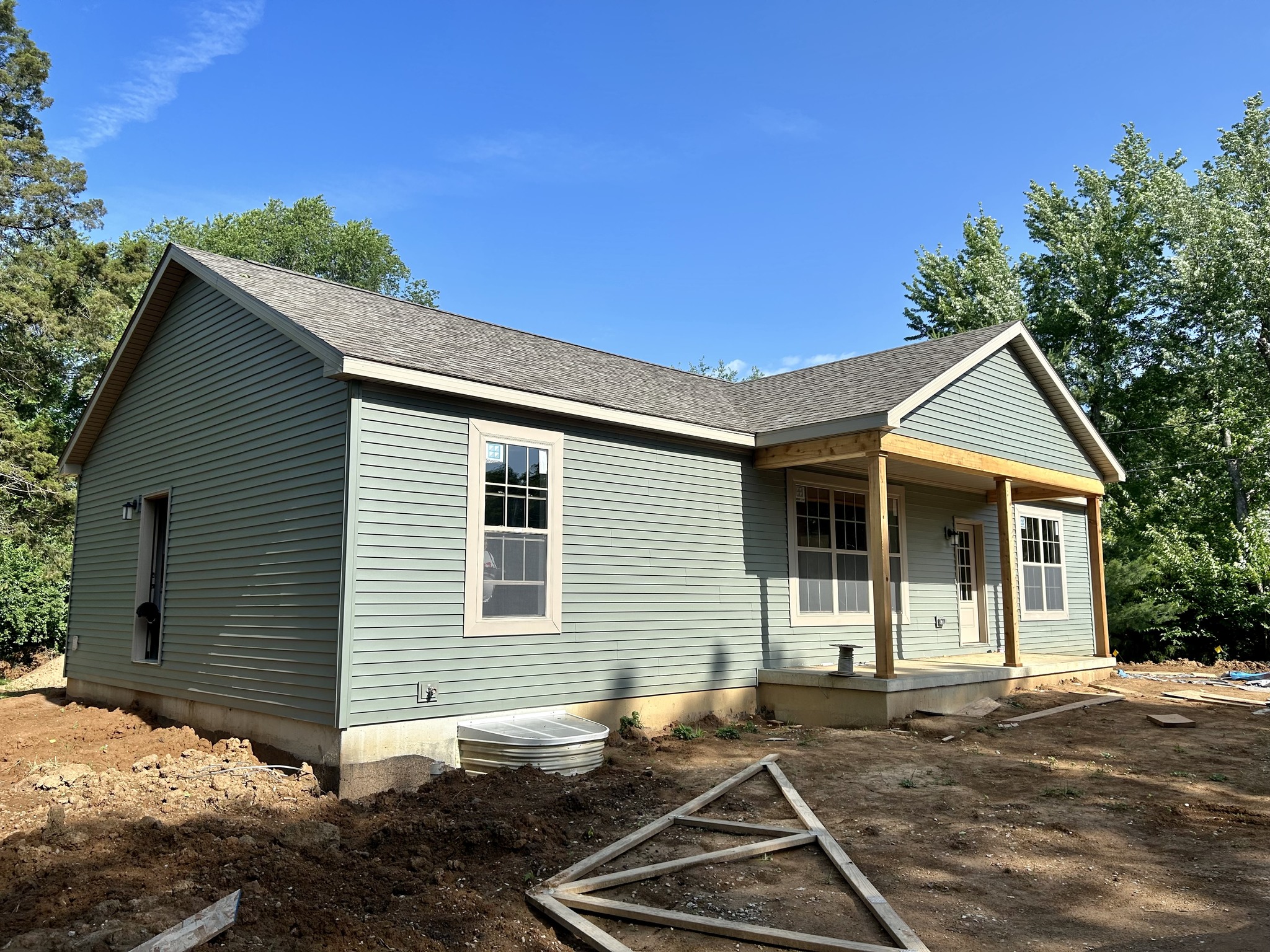 Small light green house under construction, with white-framed windows and a porch. Surrounded by trees, under a clear blue sky. Earthy ground in front.