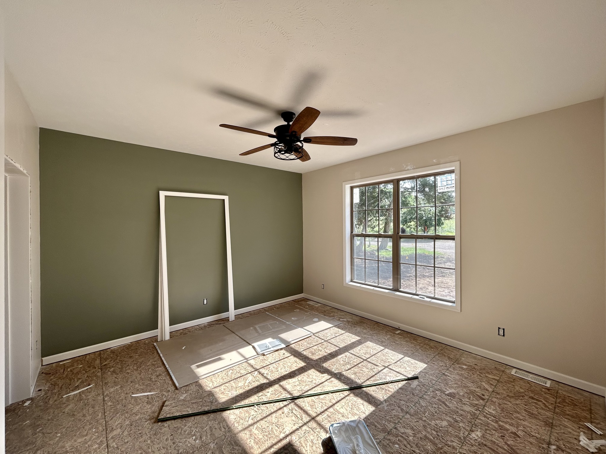 Sunlit, unfinished room with a green accent wall, large window, and textured ceiling. A fan hangs overhead, conveying a sense of renovation.
