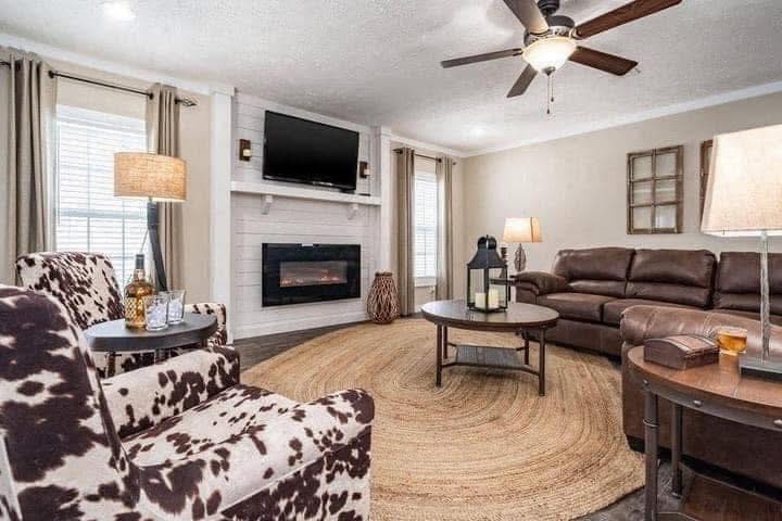 Cozy living room with a warm atmosphere. Features brown leather sofa, cow-print chairs, round jute rug, wall-mounted TV, and electric fireplace.