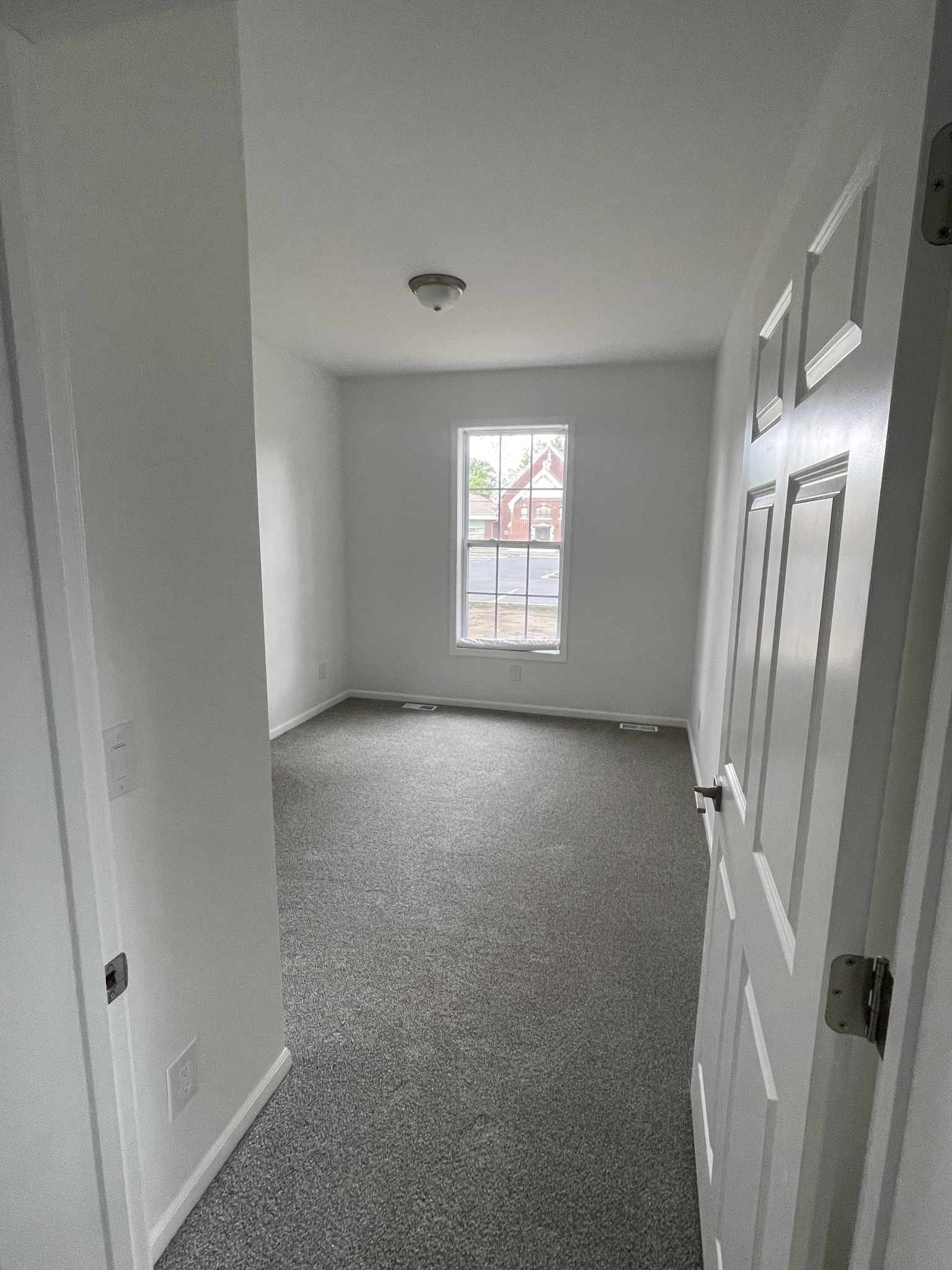 Small, empty room with white walls and gray carpet. A single window with vertical bars lets in natural light, revealing an exterior red house.