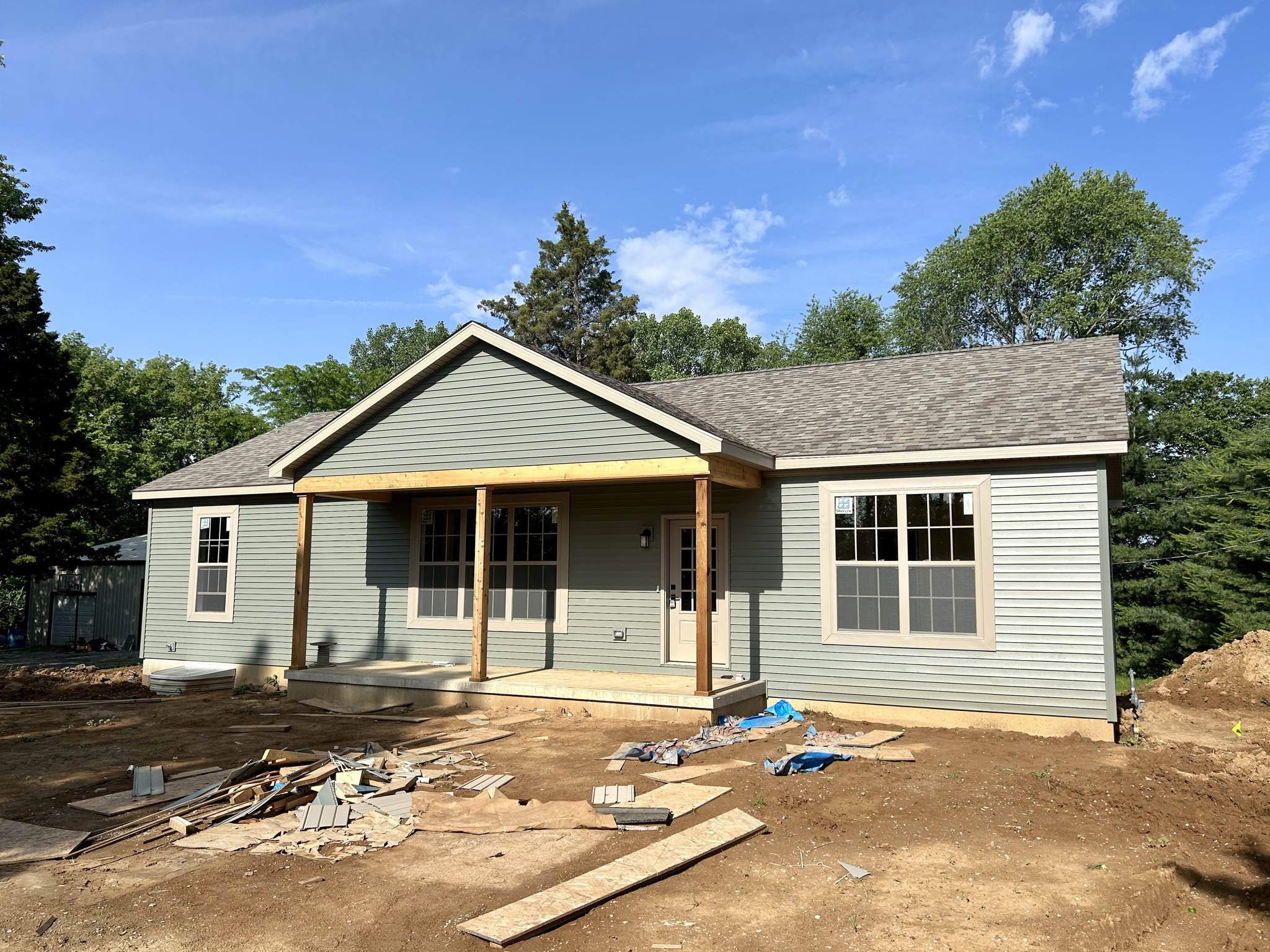Single-story gray house under construction with a small porch and unfinished front yard. Clear blue sky and lush green trees in the background.