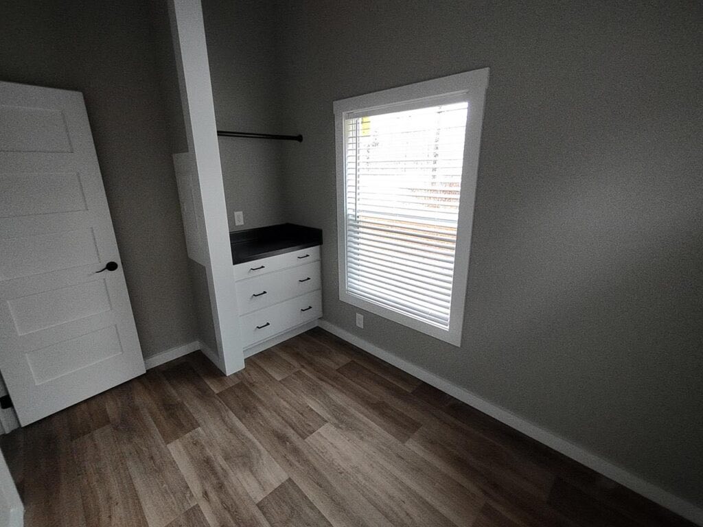 A small, empty room with light wood flooring and gray walls. It features a closet area with white drawers and a shelf, plus a window with blinds.