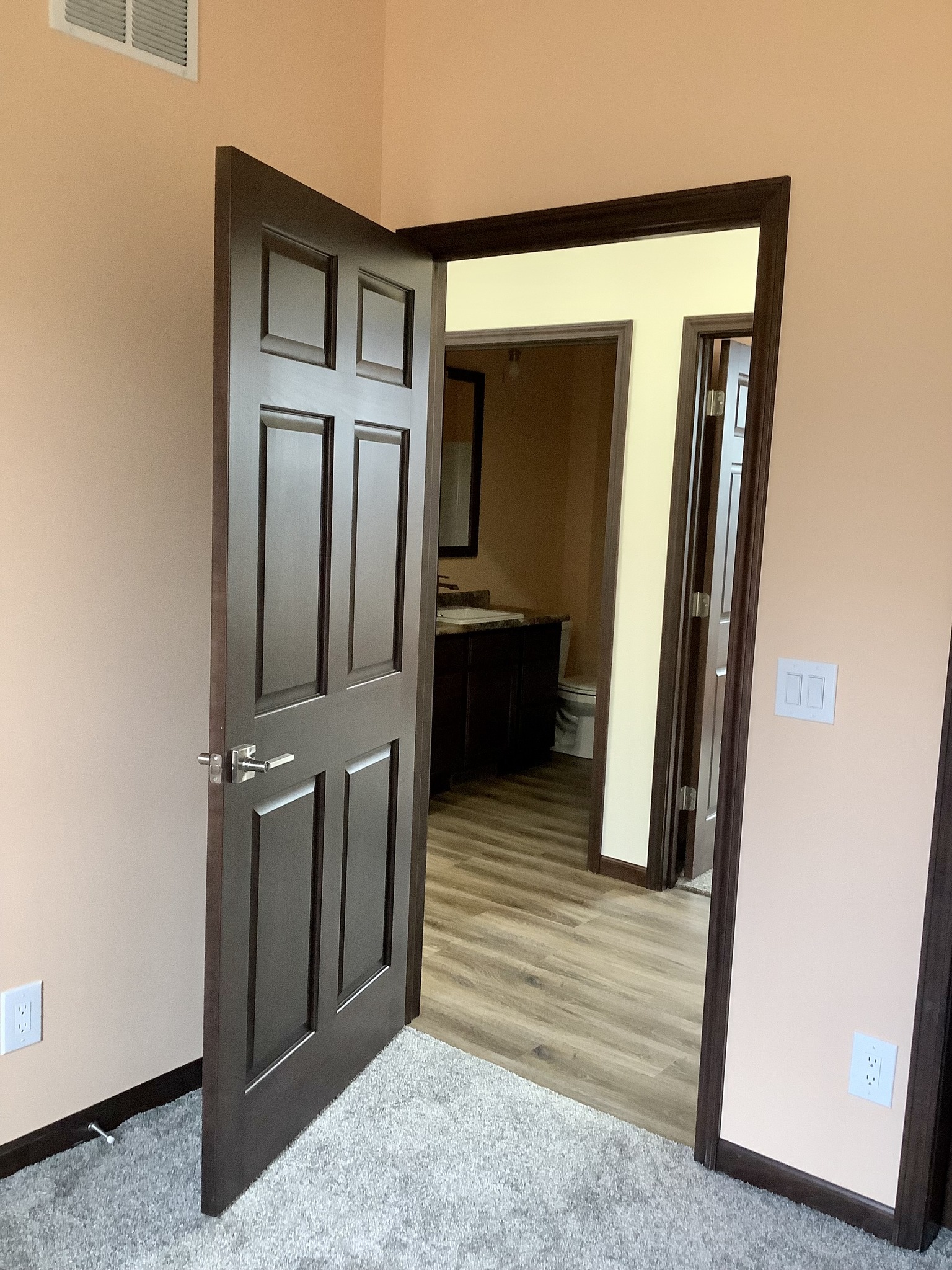 Dark wooden door ajar, opening to a room with light yellow walls, hardwood floor, and visible vanity in bathroom. Cozy, warm interior design.