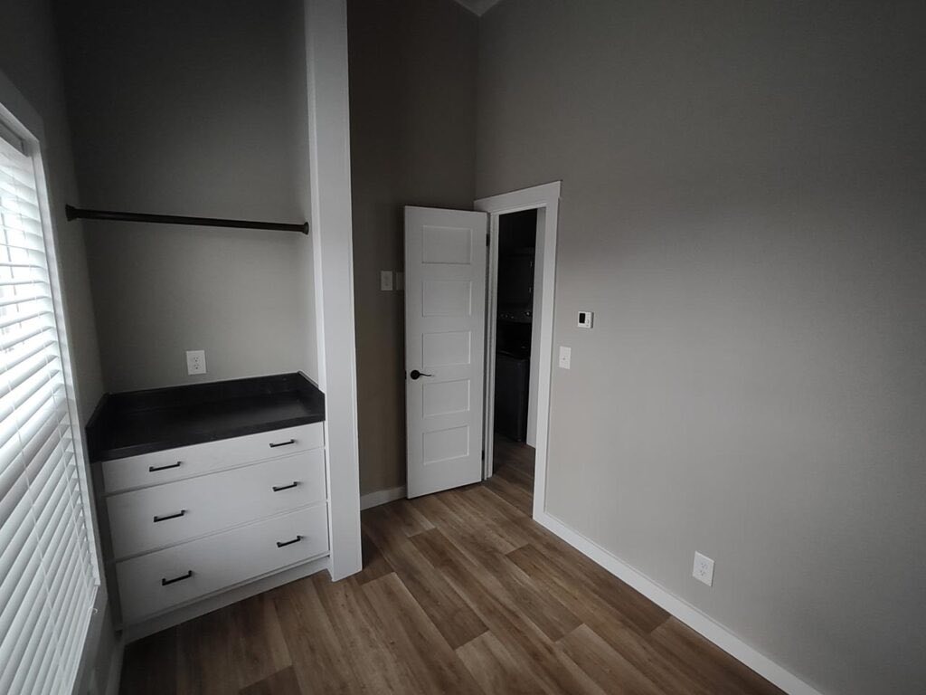 Small, empty room with light gray walls and wood flooring. Features an open white door, a closet with black countertop, white drawers, and a window with blinds.