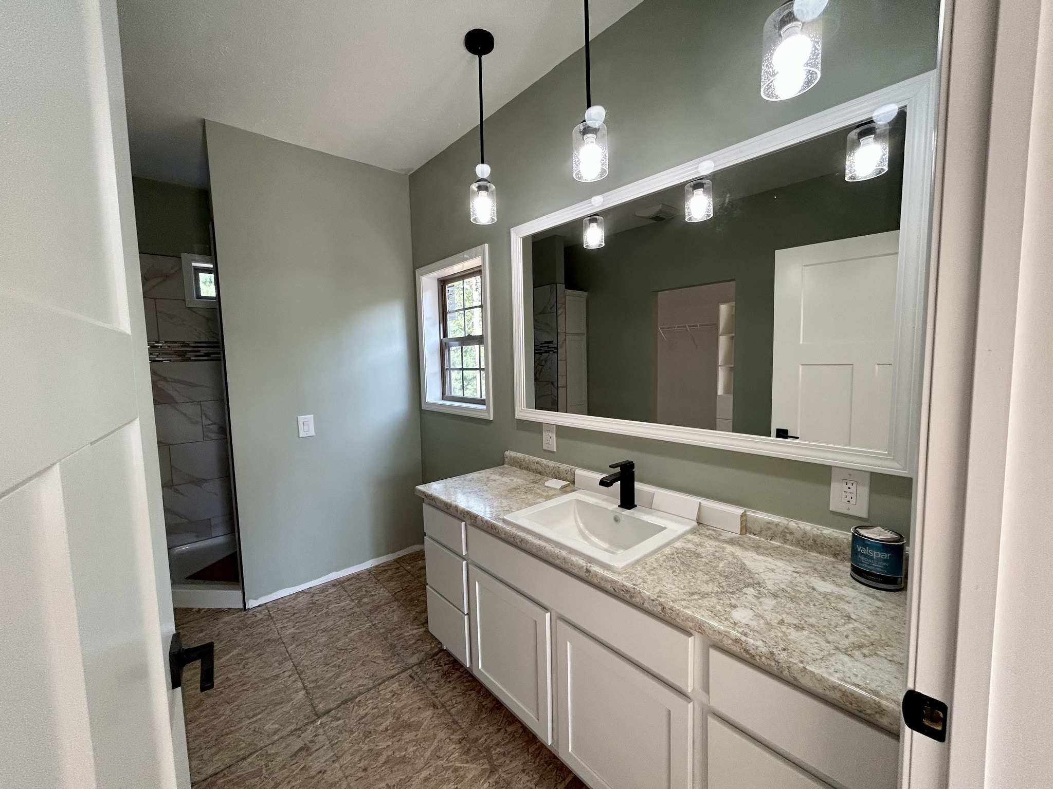 Modern bathroom with large mirror, three pendant lights, and a granite countertop. Sage green walls create a calm atmosphere. Visible walk-in shower.