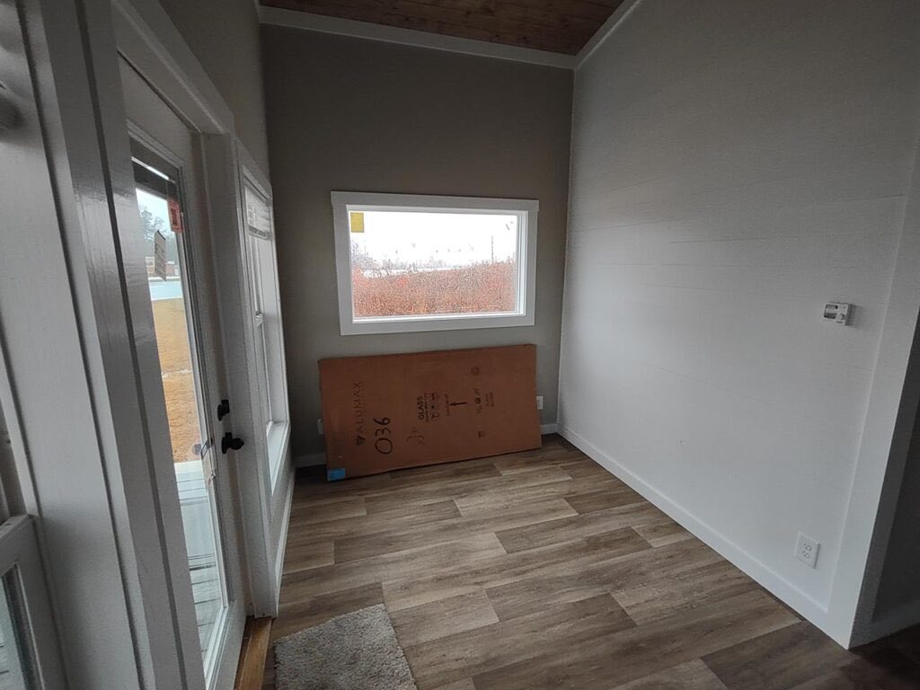 Small, empty room with a large window, wood laminate flooring, and a cardboard box against the wall. The scene appears cool and minimalist.