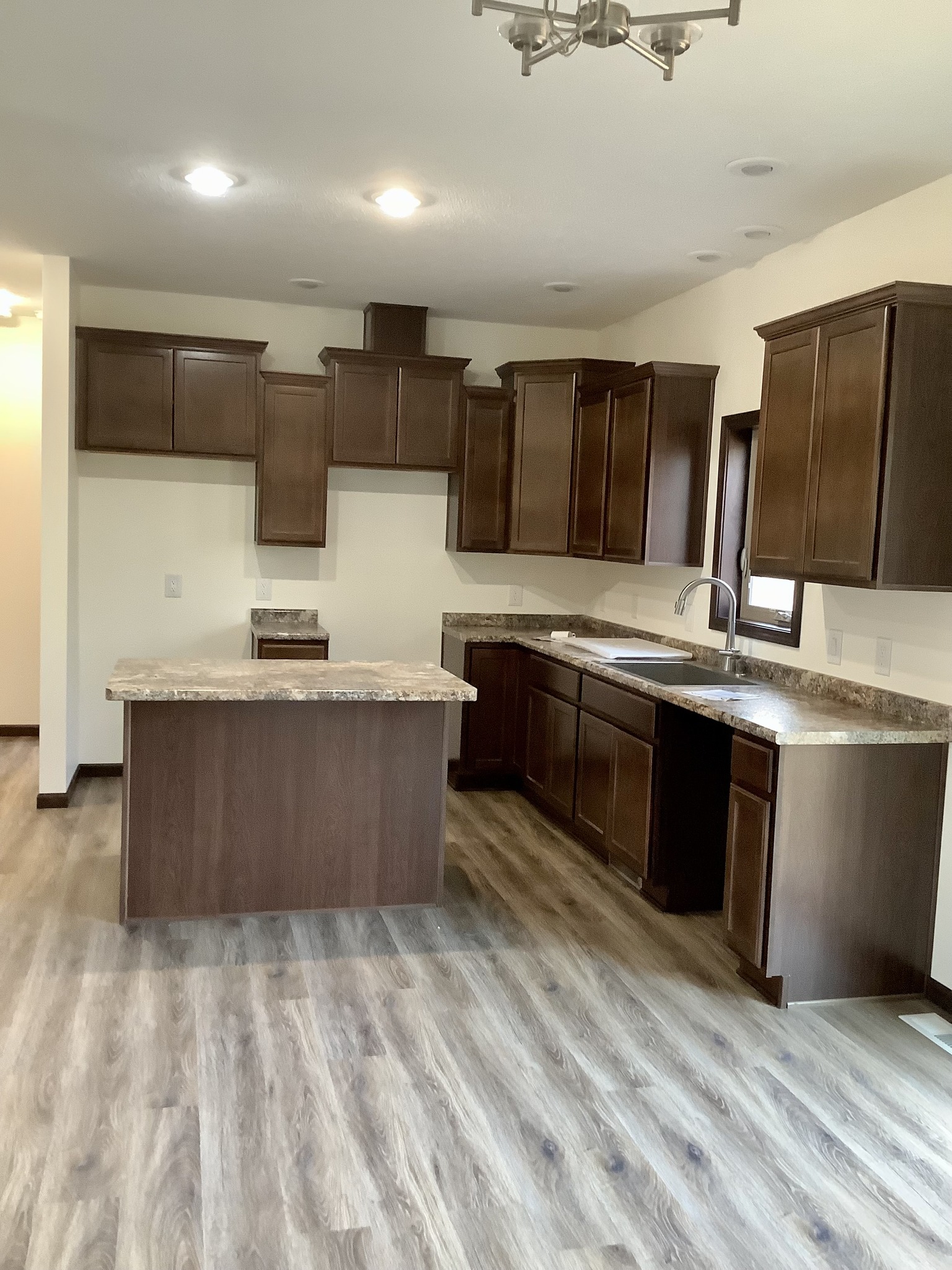 Modern kitchen with dark brown cabinets, granite countertops, and wood-look flooring. Central island under bright ceiling lights, creating a clean, warm atmosphere.