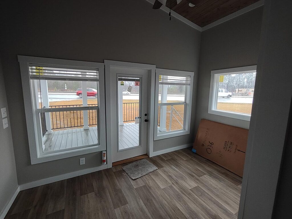 Cozy room with grey walls features large windows and a glass door, offering a view of a porch and street. Empty space has a calm, open atmosphere.