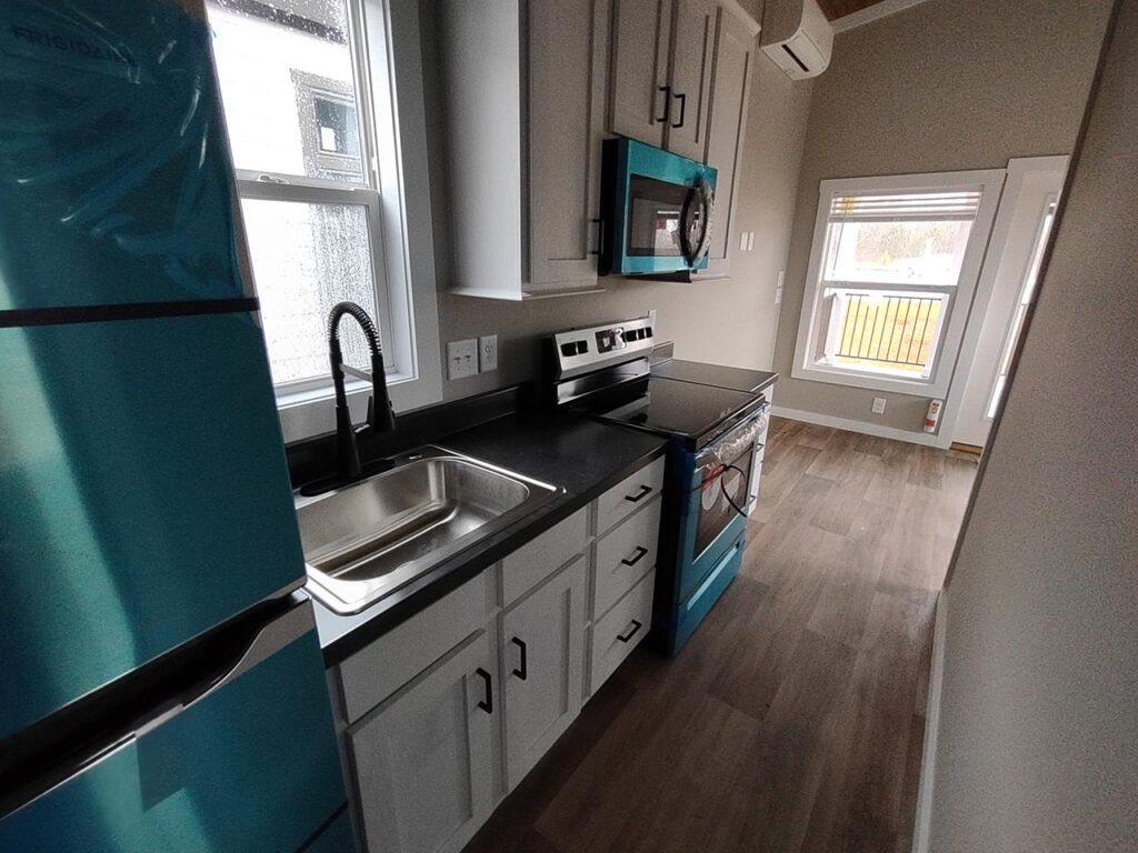Compact kitchen with gray cabinets, black countertops, and a stainless steel sink. Features a teal fridge, microwave, and stove, with a cozy, bright ambiance.