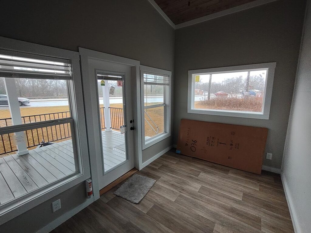A small, empty room with light brown wood flooring, large windows, and a glass door opening to a porch. A cardboard box rests against the wall, suggesting moving or renovation.