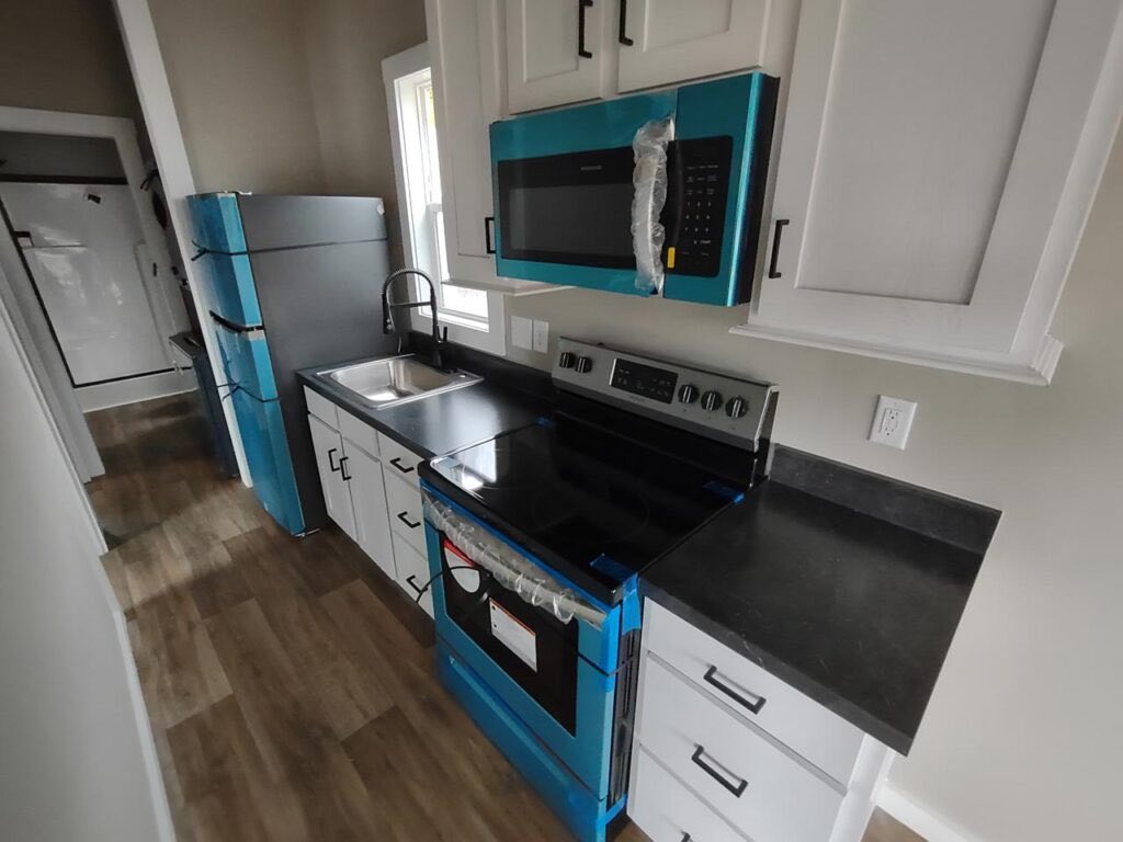 Compact, modern kitchen with wood floors, featuring newly installed black countertops, white cabinets, and appliances wrapped in blue protective film.