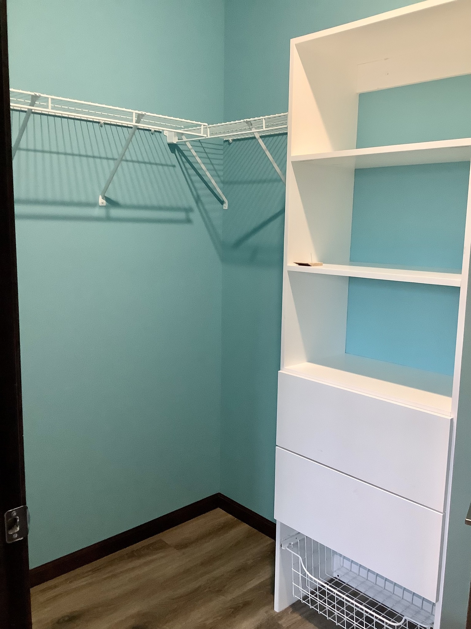 A small, teal-walled closet features white wire shelves and a modern white shelving unit with drawers. The wood floor adds warmth to the clean space.