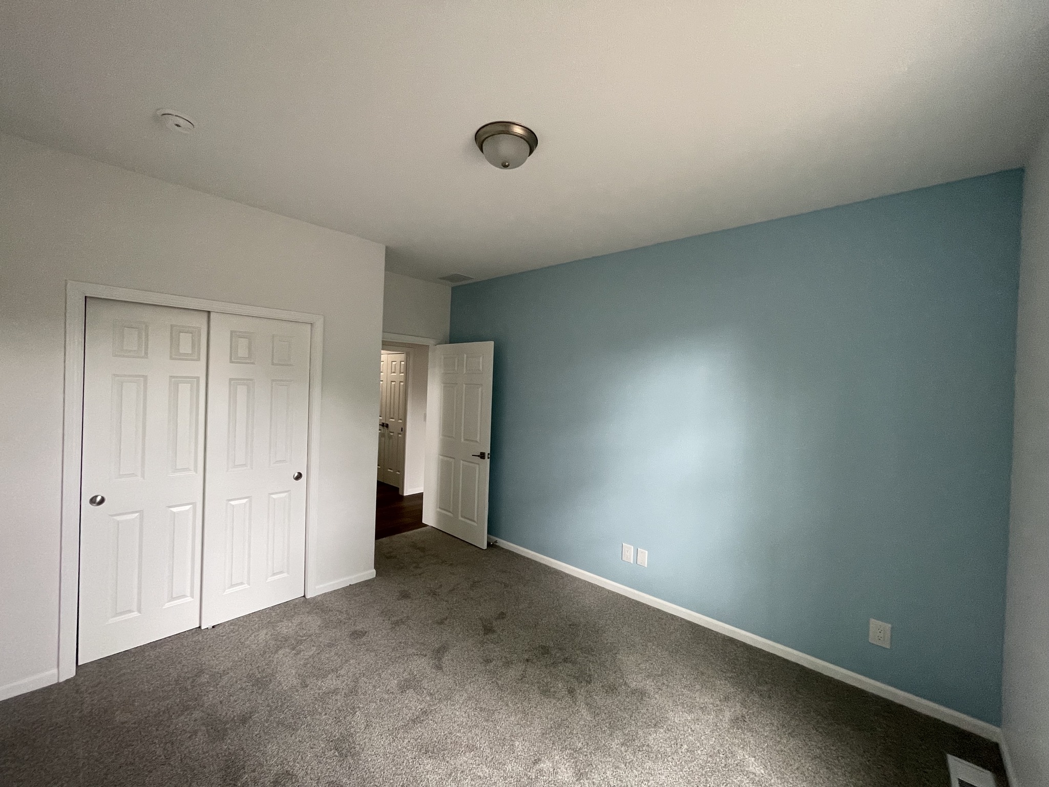 Empty room with light blue and white walls, gray carpet, and a closed closet door on the left. A white door is slightly open, revealing a hallway.