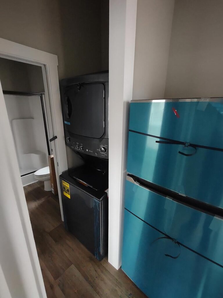 Compact laundry area with a gray stacked washer and dryer beside a blue cabinet. A bathroom with a white toilet and shower is visible in the background.