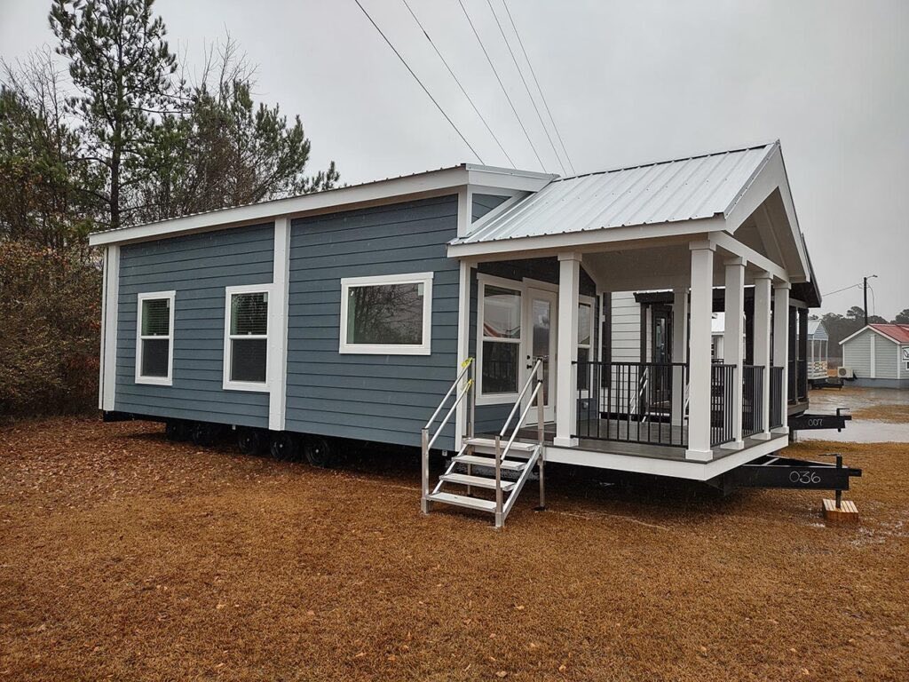 A compact, modern tiny home with blue siding and white trim is on a grassy lot. It has a small porch, large windows, and a metal roof on a rainy day.