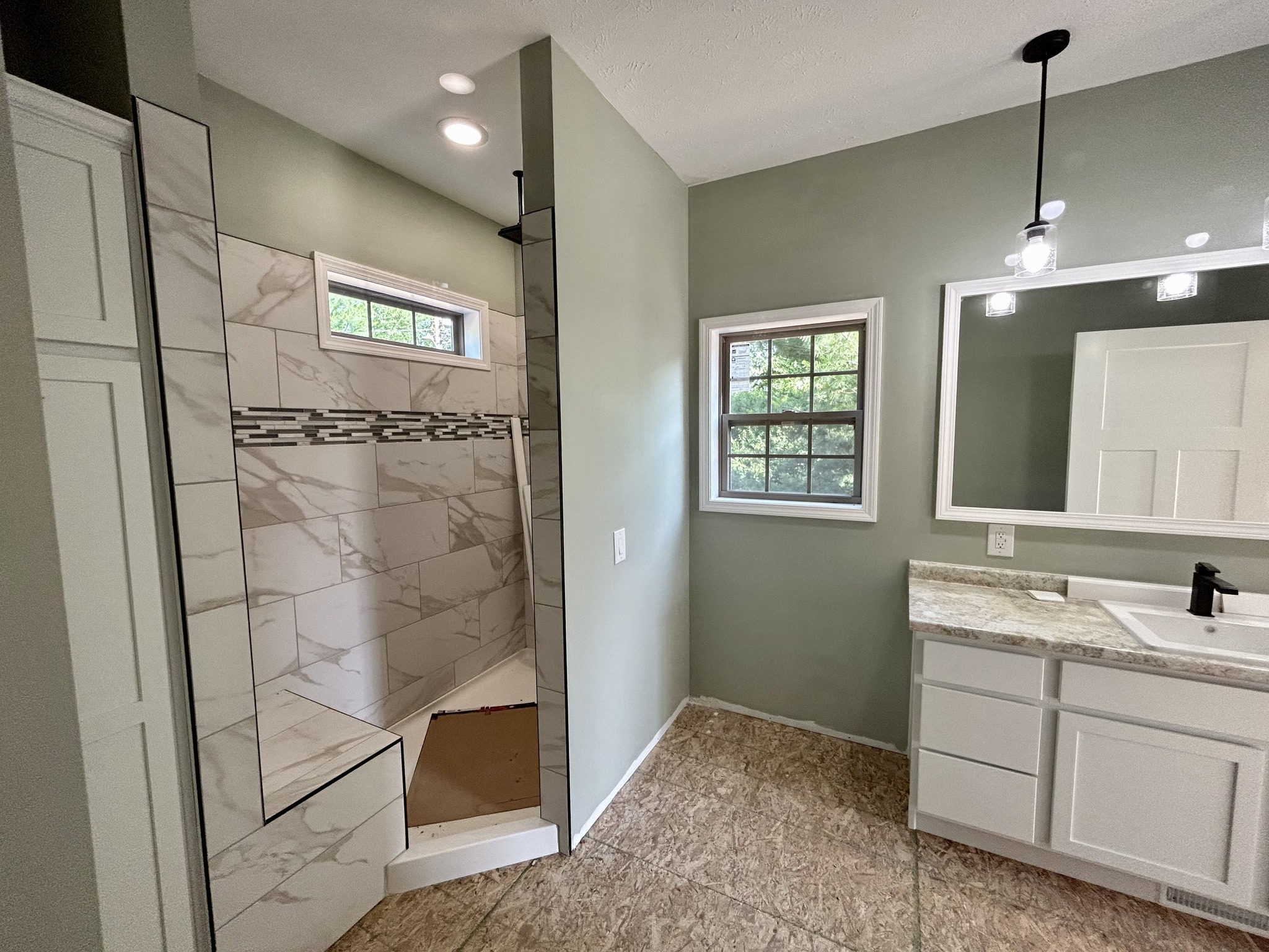 Modern bathroom with green walls and tiled walk-in shower featuring a marble pattern and mosaic accent. A vanity with a large mirror and pendant light is visible.