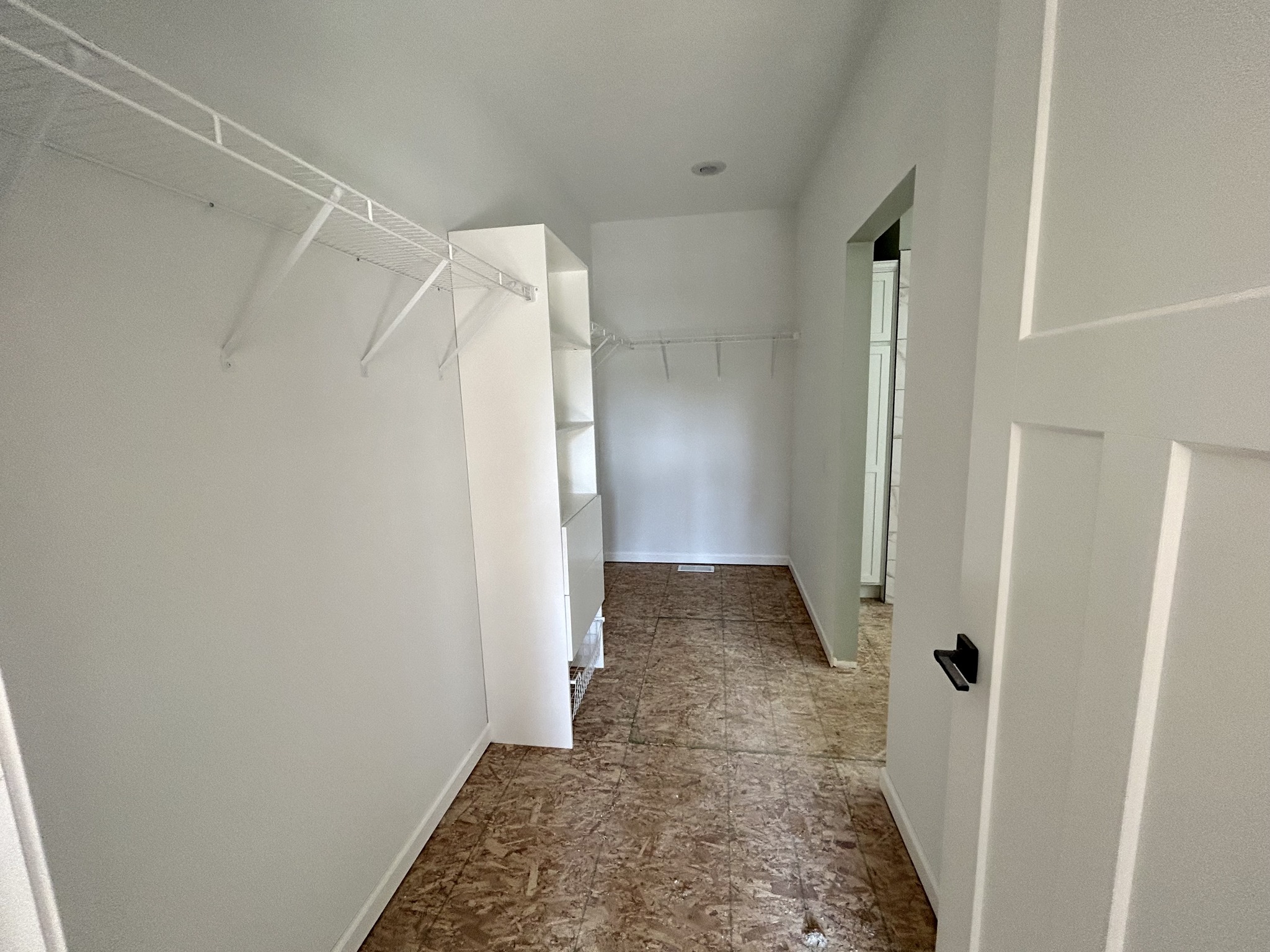 Empty walk-in closet with white walls and wire shelving on both sides. It has an open doorway, plywood flooring, and a clean, minimalistic feel.
