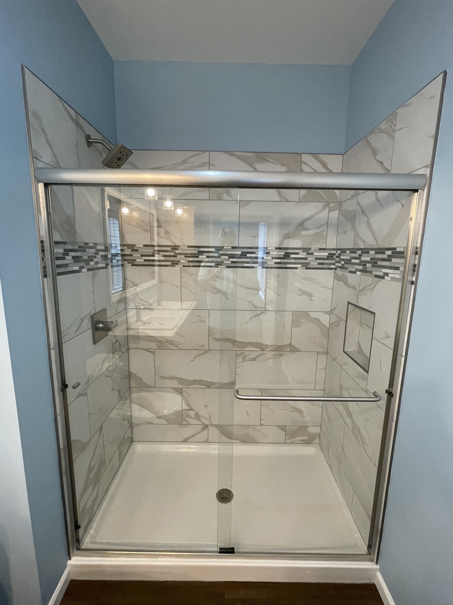 Modern shower with glass door, featuring white marble tiles and a decorative mosaic stripe. It includes a metal grab bar and built-in shelf. Elegant and clean design.