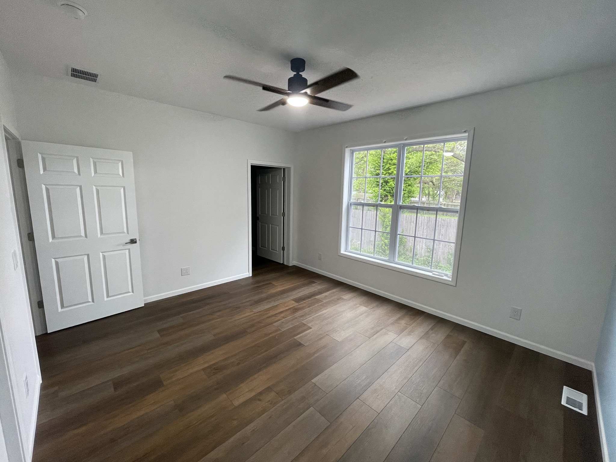 A bright, empty room with white walls and dark wood flooring. A ceiling fan with a light is on. A large window shows a green view outside.