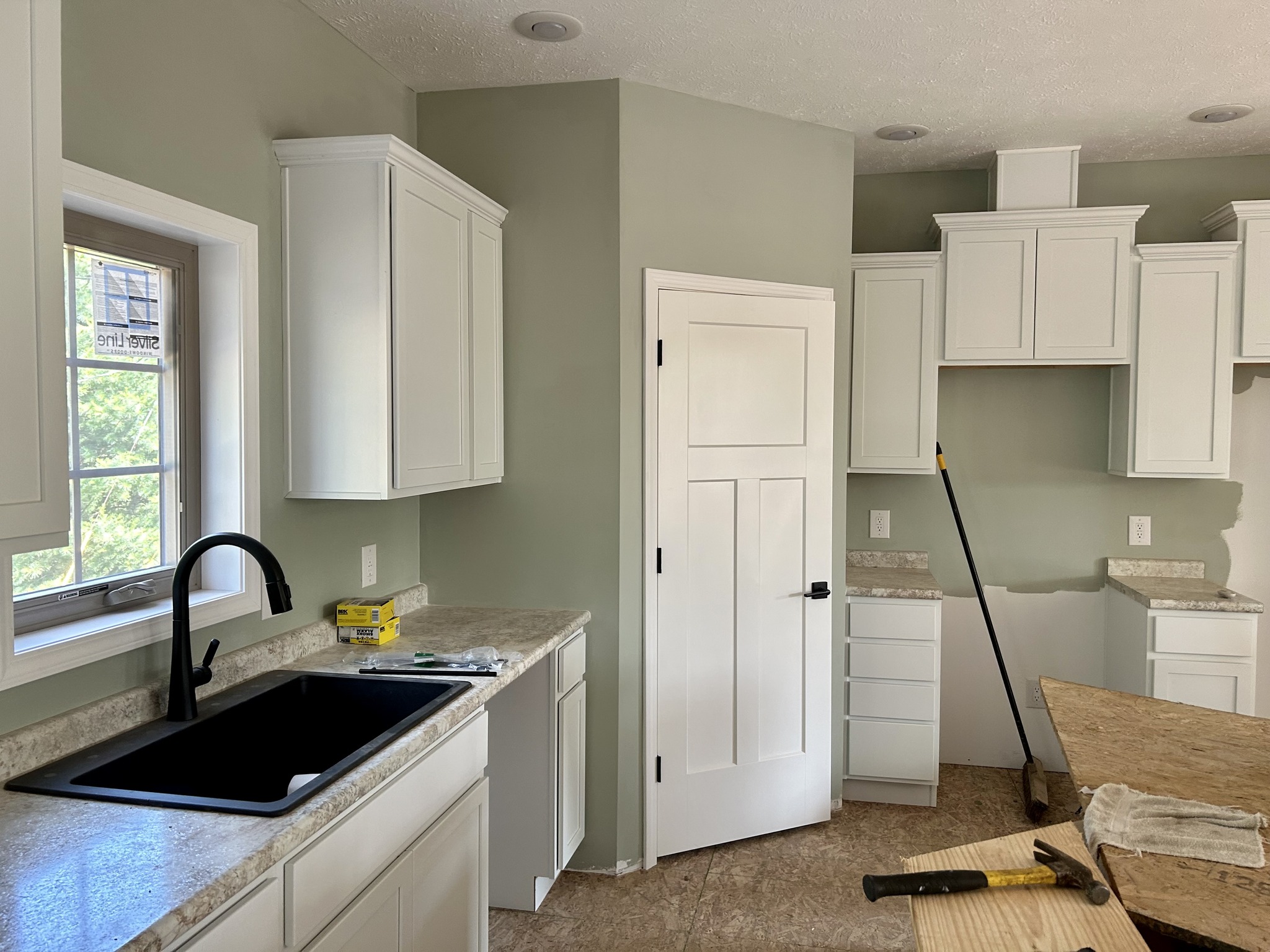 Modern kitchen under renovation; white cabinets, light green walls, black sink, and granite counters. Tools on the countertop suggest ongoing work.
