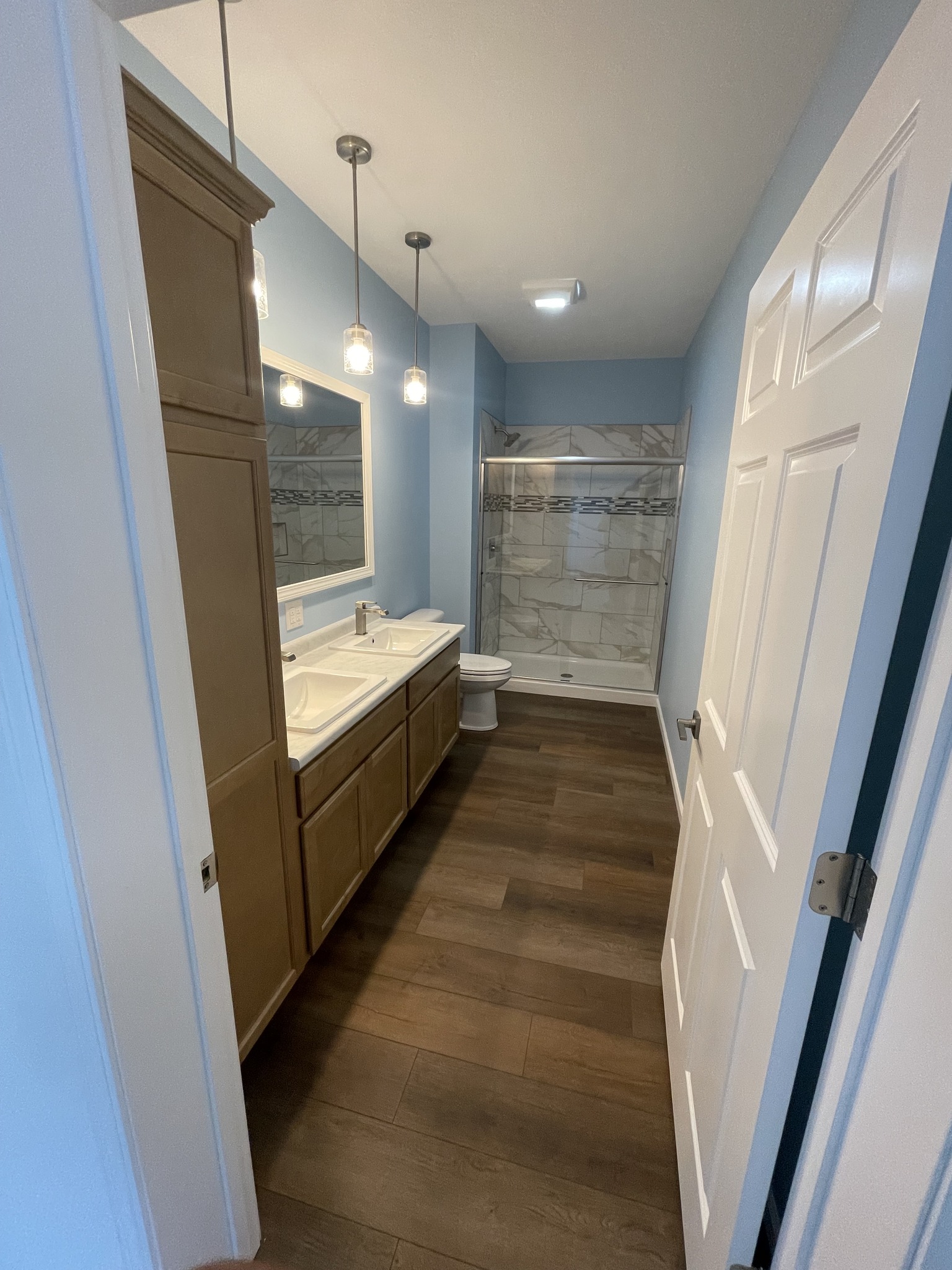Modern bathroom with light blue walls, wood flooring, and three pendant lights. Features a double sink vanity, marble shower, and toilet. Calm ambiance.
