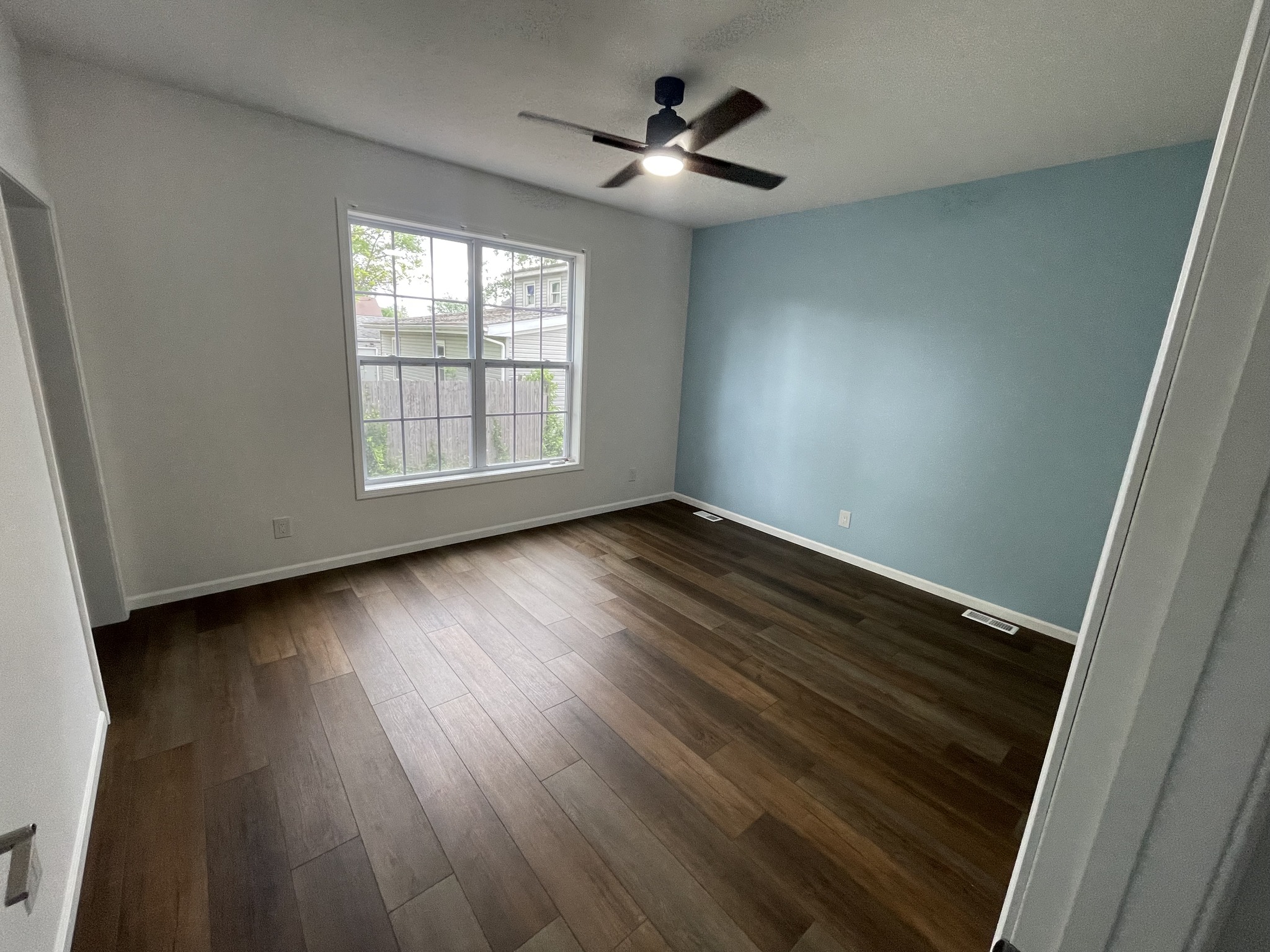 Empty room with wooden floors and a large window on the left, light blue accent wall on the right, and ceiling fan. Bright, airy atmosphere.