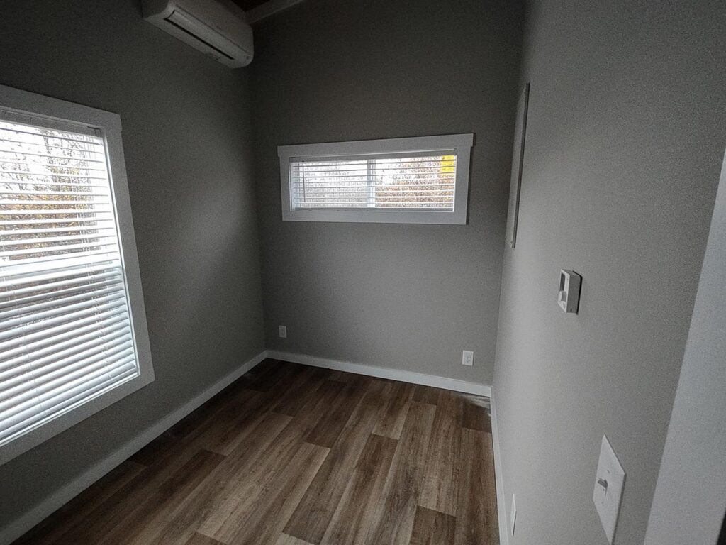 A small, empty room with gray walls and wooden flooring. Two horizontal windows with blinds allow natural light. An air conditioning unit is mounted above.