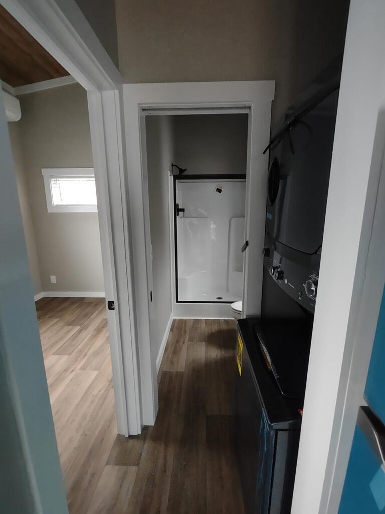A compact room featuring a stackable washer and dryer on the right. A bright, narrow hallway leads to a white shower stall, with wood laminate flooring throughout.