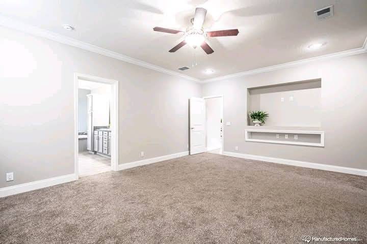 Spacious beige living room with carpeted floor, ceiling fan, and built-in shelving. Open door reveals a bathroom with dual sinks. Bright and inviting.