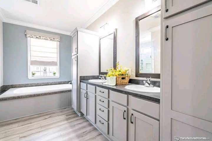 Bright bathroom with gray cabinets and dual sinks with mirrors above. A window above a bathtub lets in natural light. Yellow flowers add a warm touch.