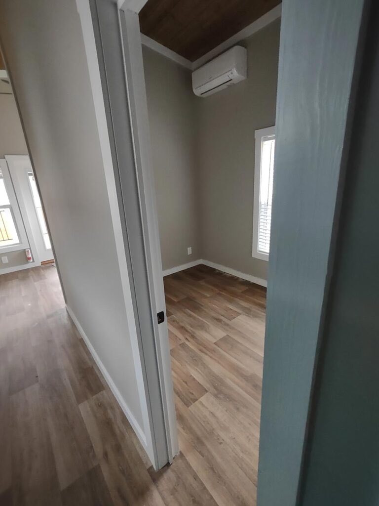 Narrow hallway leading to a small room with light wood flooring, gray walls, and a window. An air conditioner is mounted on the wall, creating a minimalist atmosphere.