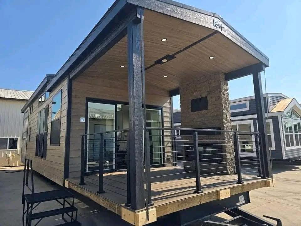 Modern tiny house with large windows and covered porch. Features a wood and stone exterior, black railings, and a bright, open interior.