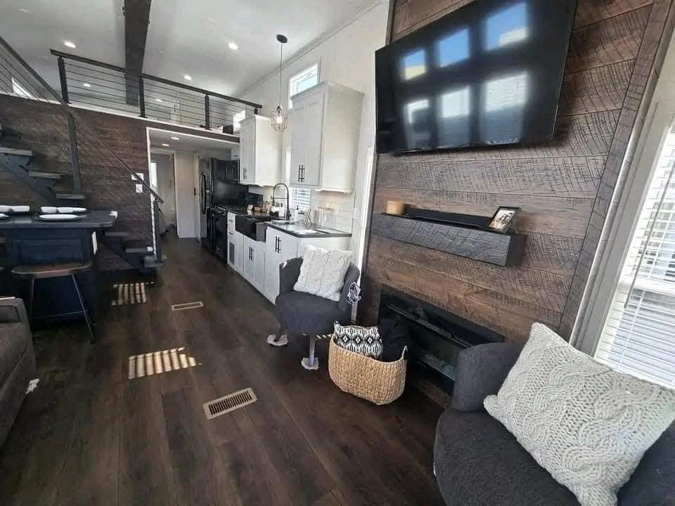 Modern tiny home interior with dark wood floors, a TV on rustic wood paneling, kitchen with white cabinets, loft, cozy chairs, and natural light.