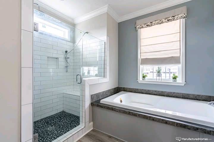 Modern bathroom with a glass-enclosed shower featuring light tiles and dark pebble flooring. Adjacent to the shower is a large bathtub under a window with a gray and white design, creating a clean and serene atmosphere.