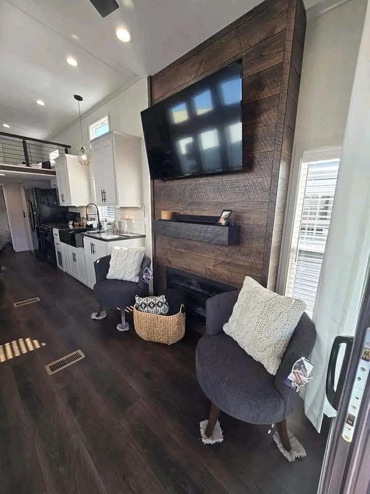Modern living area with dark hardwood floors, featuring two gray cushioned chairs with cream pillows. A wall-mounted TV sits above a rustic wood panel fireplace. White kitchen cabinets and a black countertop are visible. Bright, airy ambiance.