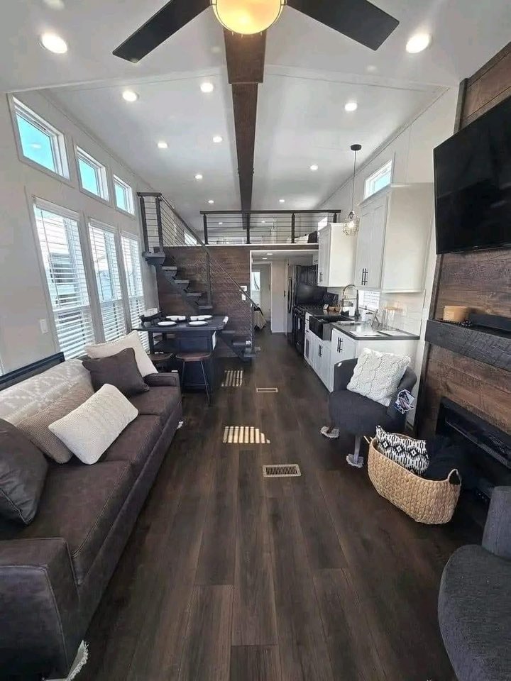 Modern tiny home interior with dark wood floors, cozy seating, and a loft. Bright windows and a sleek kitchen create a spacious, inviting feel.