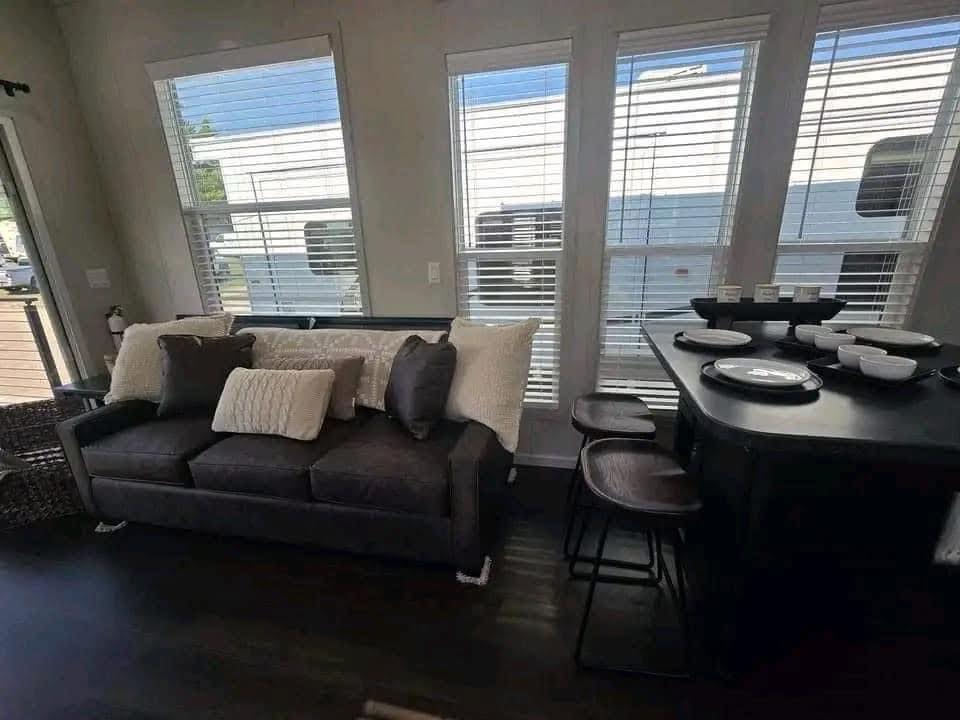 Modern living area with dark sofa adorned with cream and brown pillows, facing a dining counter with bar stools and dishware. Large windows offer natural light.