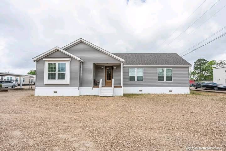 Gray manufactured home with white trim sits on a gravel lot under a cloudy sky. Features a small porch with a chair. Surrounding trees add greenery.
