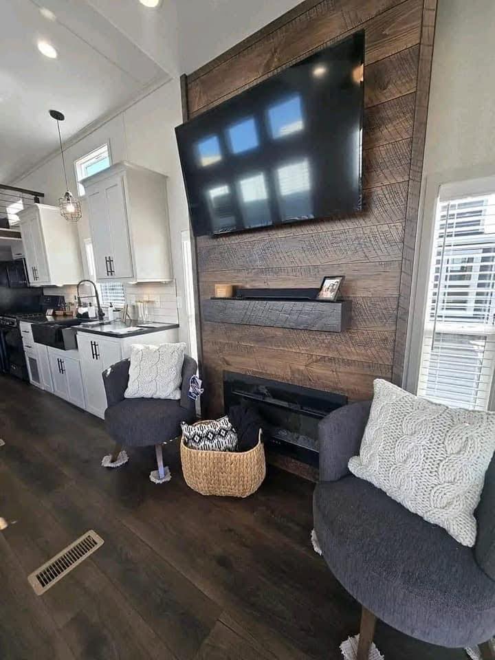 Modern living room with a wall-mounted TV above a wooden panel, flanked by two gray chairs with white cushions. Cozy ambiance with a woven basket.
