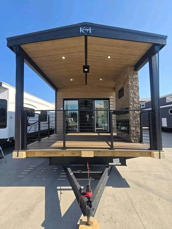 Modern tiny home on wheels with a wooden porch and stone accents. Large glass doors under a sloped roof. Bright, sunny day in a parking lot setting.
