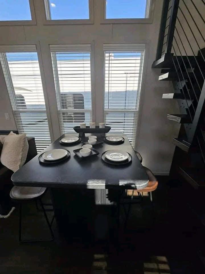 Dining table set for four with black plates and white cups, near large windows with blinds. Modern staircase to the right, bright daylight fills the room.