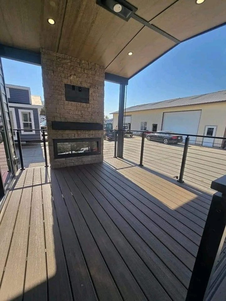 Covered patio with a stone fireplace and glass firebox. Wood panel ceiling, dark flooring, black railing, and nearby modern buildings under clear sky. Relaxing ambiance.