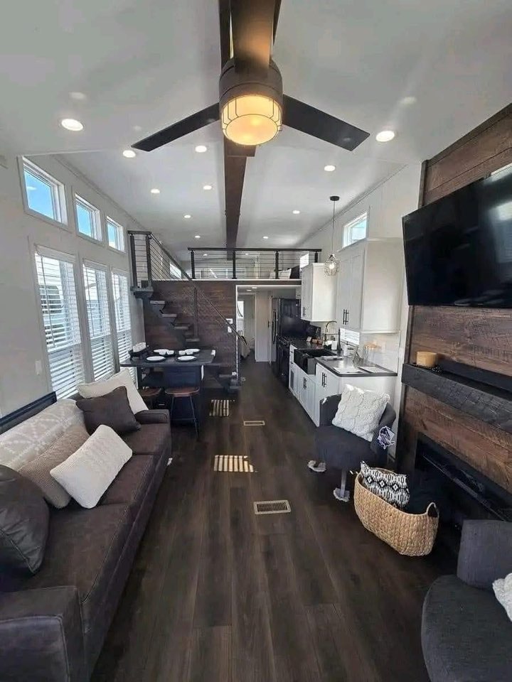 Modern tiny home interior with high ceiling and dark wood floors. Features a sofa, staircase, loft, kitchen, and large windows. Cozy and stylish.